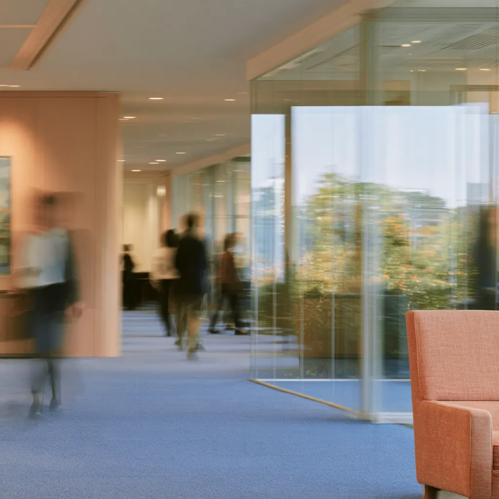 An office hall with glass walls, people blurred mid-step along the corridor, an orange chair in the foreground and trees beyond the glass