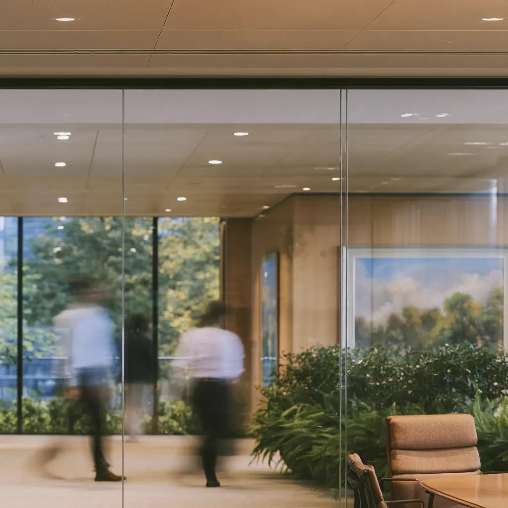 A glass-walled conference room in a law firm, a framed landscape painting on the wood wall behind it, blurred figures walking past the glass