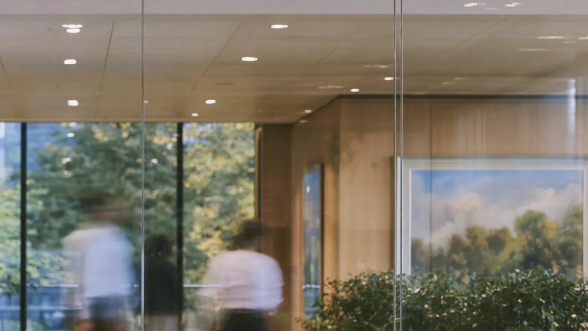 A glass-walled conference room off an office hall, people passing in a blur on the other side of the glass